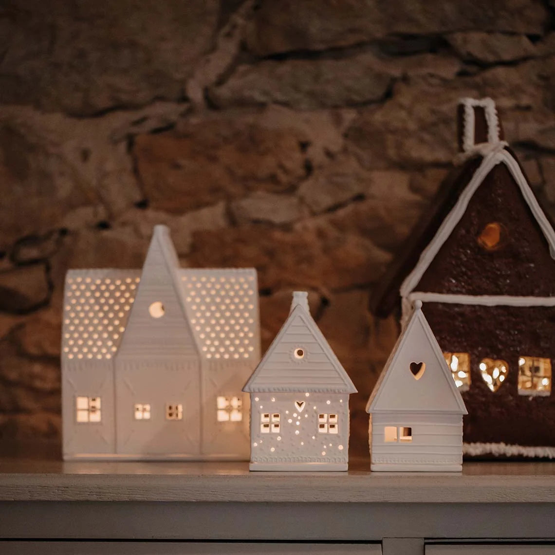 Collection of white tealight house candle holders shaped like gingerbread houses on a wood drawer unit against a rock wall background