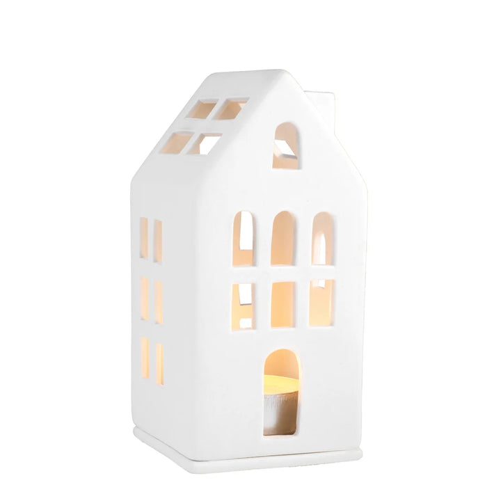 A white ceramic house-shaped candle holder with arched and rectangular cutouts, illuminated from within by a warm yellow light, set against a plain white background.