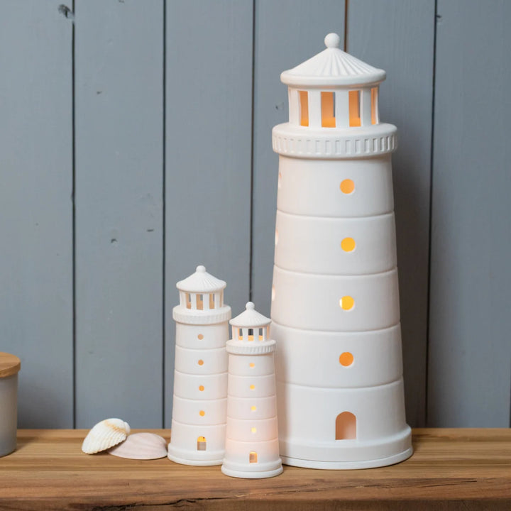 Collection of white tealight house lighthouse candle holders with lit candles on a timber shelf against a grey wall