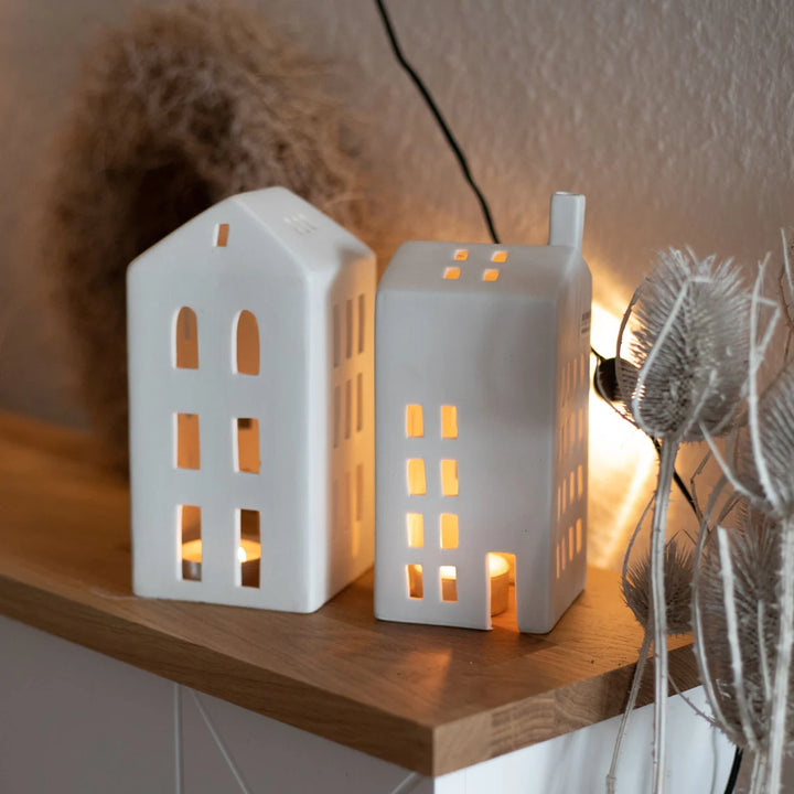 Two white ceramic house-shaped tealight candle houses on a wooden surface with a warm glow.
