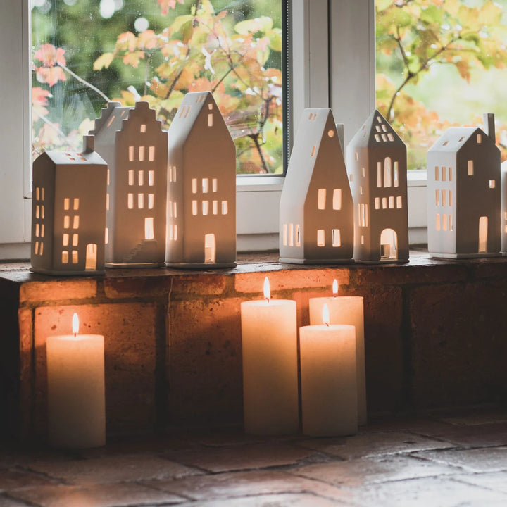 A set of ceramic miniature house-shaped candle holders with glowing warm lights, displayed on a brick windowsill with real pillar candles in front and an outdoor window view in the background.