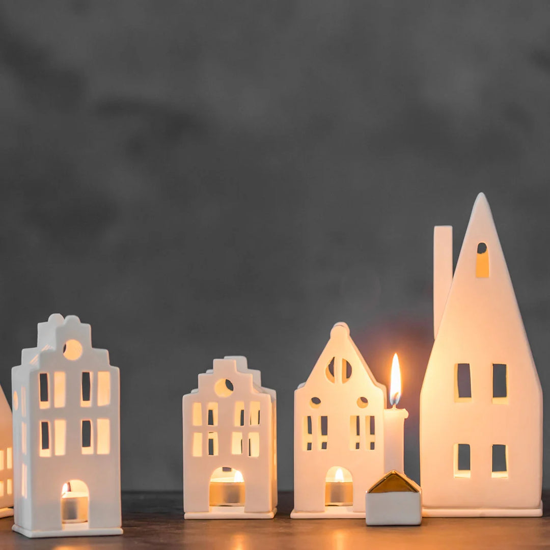 Collection of four white porcelain tealight house candle holders with lit candles on a black surface with a grey background