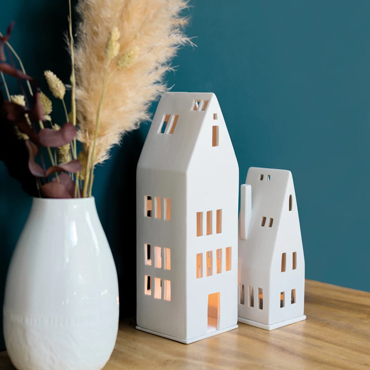 Two white porcelain tealight house candle holders in front of a teal wall on a wood table