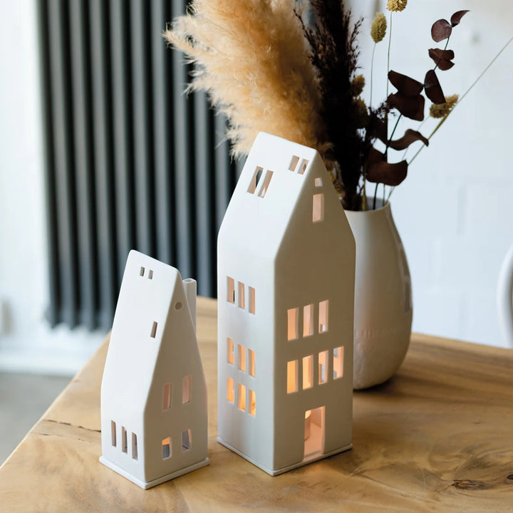 Two porcelain tealight house candle holders on a wood table with a dried flower arrangement