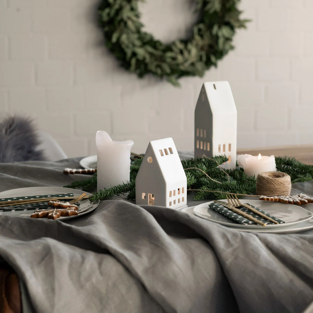 Styled table with grey tablecloth, plates with gold cutlery, white tealight house candle holders and a green wreath in the background