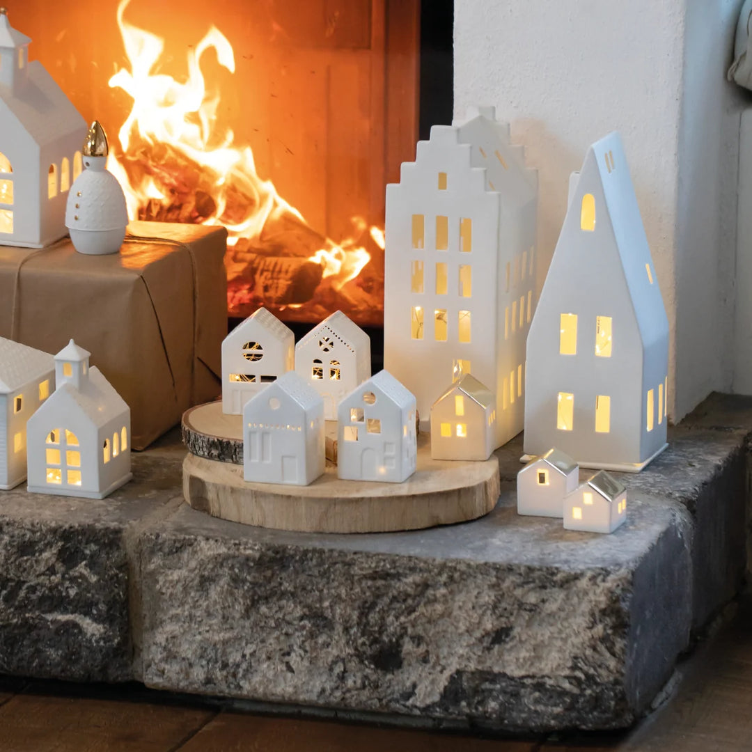 Collection of white porcelain tealight house candle holders wtih lit candles arranged in front of a fireplace