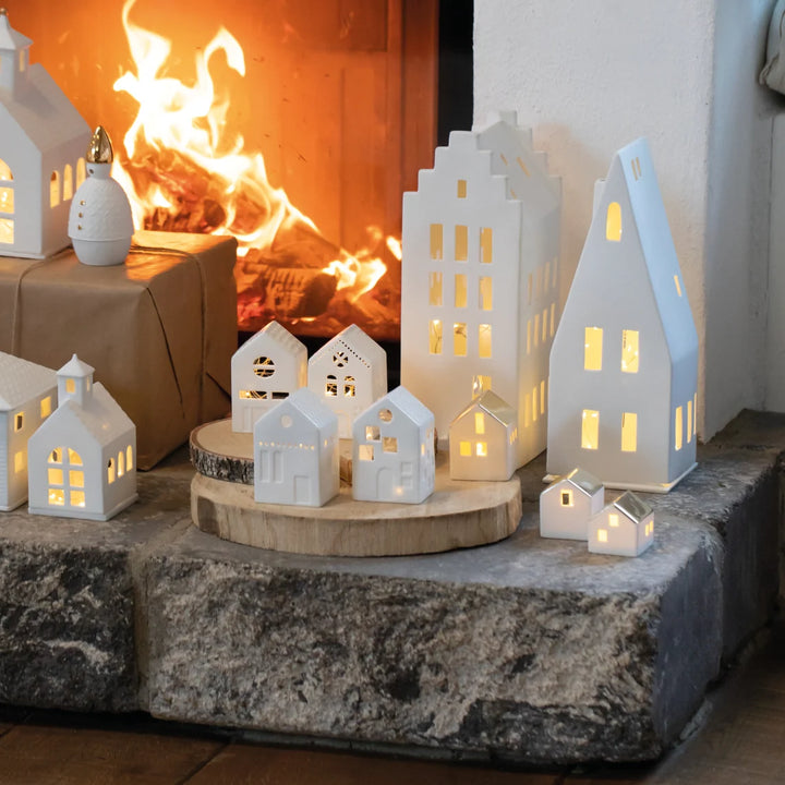 Collection of white porcelain tealight house candle holders wtih lit candles arranged in front of a fireplace