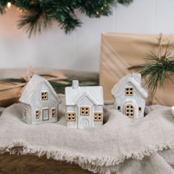 Three small white ceramic house-shaped candle holders with warm tan window details, arranged on a beige fringed textile with wrapped gifts and evergreen branches in the background.