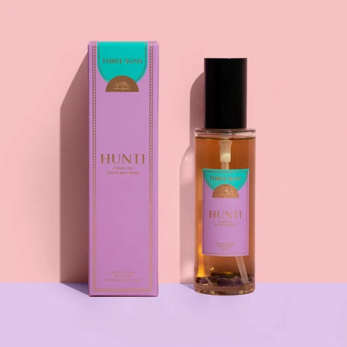 A bottle of THREE SUNS Hunti room spray next to its lavender and teal packaging box, set against a pastel pink and purple background.
