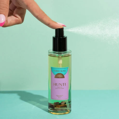 A hand with pink nails sprays a fine mist from a Three Suns Hunti room spray bottle. The bottle is filled with a light green liquid and features a black pump and a lavender label, set against a solid mint green background.