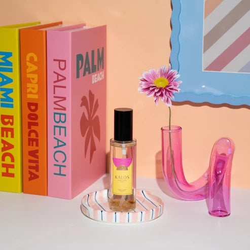 A small clear glass room spray bottle with a black cap and yellow label reading KALOS stands on a striped coaster beside a pink U-shaped vase holding a flower, with colorful books in the background against a peach wall.