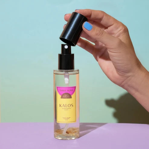 A hand with blue nail polish holds the black spray cap above a bottle of Three Suns Kalos room spray against a pastel mint and lavender background.