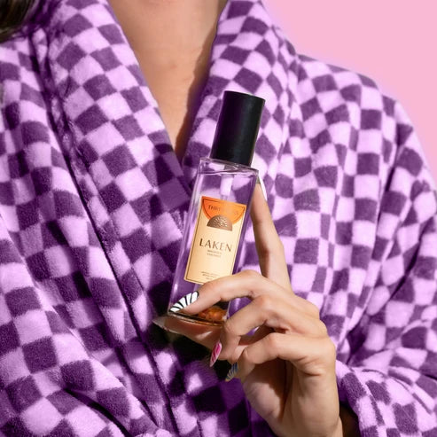 A person wearing a purple and lavender checkered bathrobe holds a bottle of Laken room spray against a solid pink background.