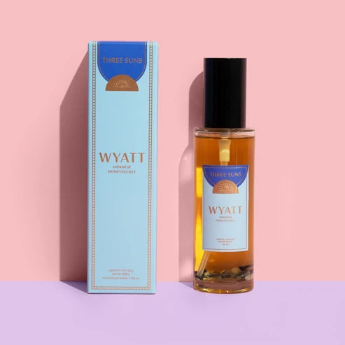 A bottle of Wyatt Japanese Honeysuckle room spray next to its light blue packaging box, set against a split pink and lavender background.