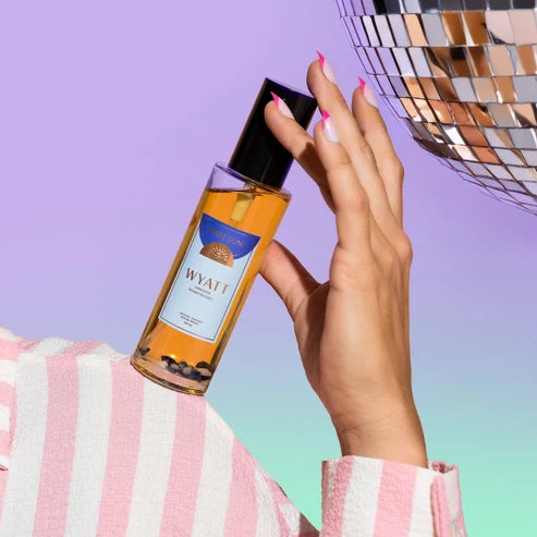 A hand with pink manicured nails holds a bottle of Wyatt room Spray against a pastel purple and mint gradient background, with a pink-and-white striped fabric and a disco ball visible.