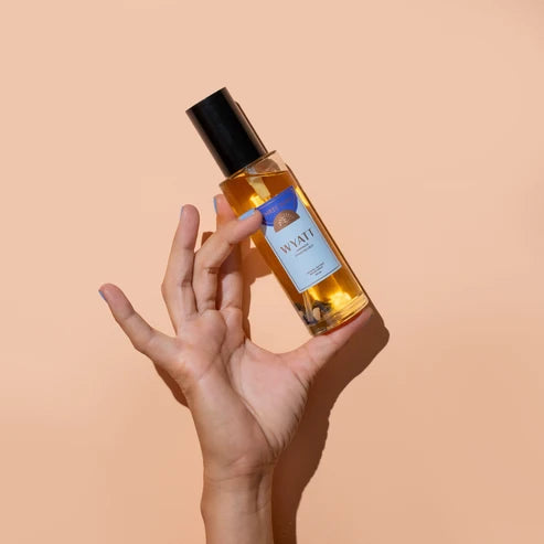 A hand holds a clear bottle of Wyatt Room Spray with golden liquid inside, a black cap, and a white label with blue accents, set against a solid peach background.