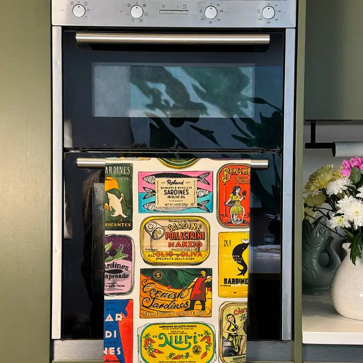 A stainless steel built-in oven with a glass door, featuring a colorful vintage-style collection of food tins and labels hanging from a horizontal handle on the oven door. The oven is set against a dark olive-green background, with a vase of flowers visible on a shelf to the right.