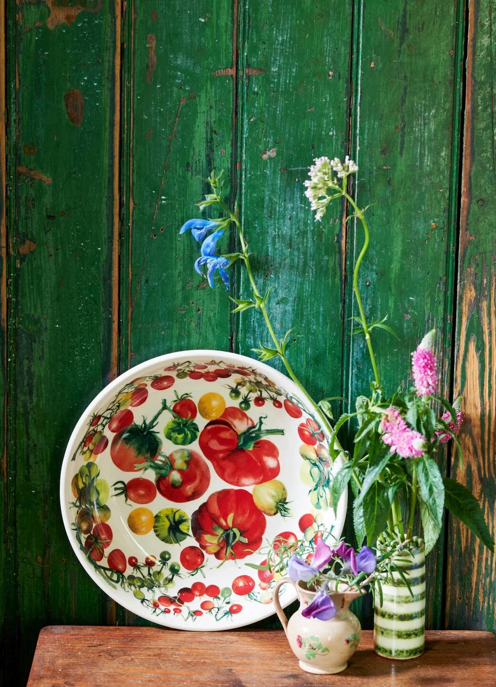 A decorative ceramic bowl with a colorful tomato print, styled on a rustic wooden table against a distressed green wooden wall, alongside small vases with flowers.