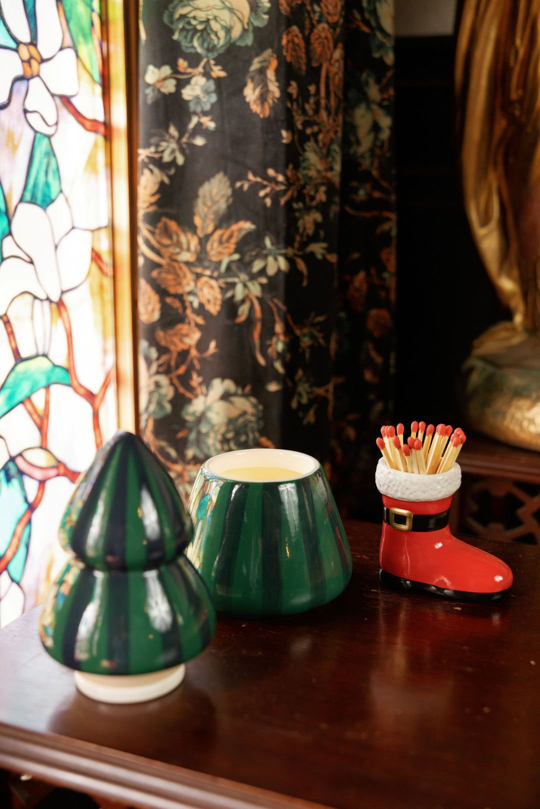 Three small decorative ceramic containers on a dark wooden table: a green Christmas tree-shaped candle holder lid, candle base with cream candle inside, and a red Santa boot-shaped match holder with red match tips. A stained-glass panel and floral-patterned curtain are visible in the background.