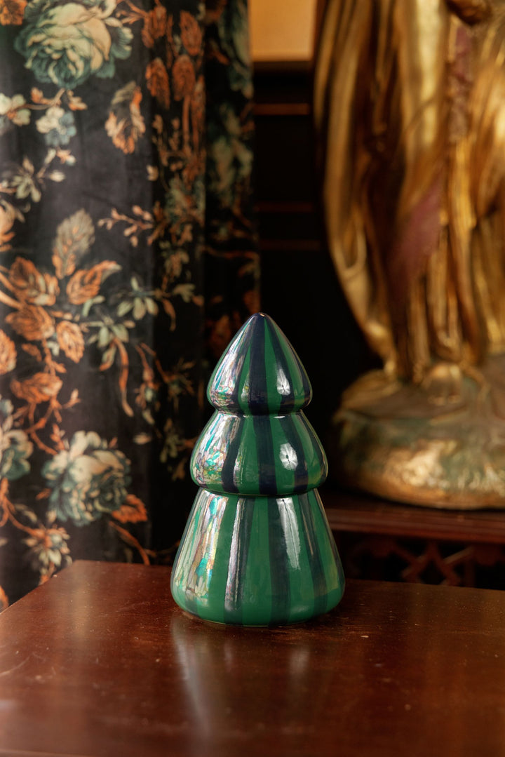 A glossy green ceramic Christmas tree-shaped candle, displayed on a dark wooden table in an indoor setting with a floral curtain and a gold-toned statue in the background.