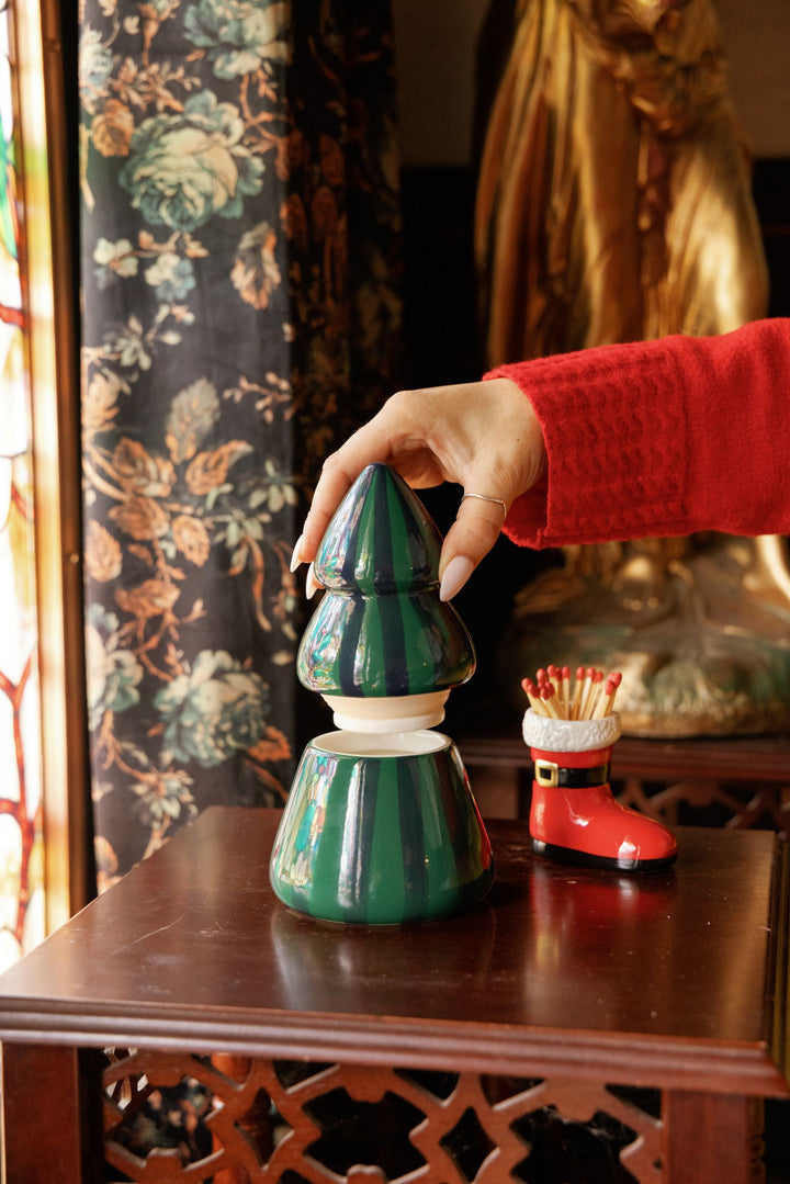 A hand wearing a red knit sweater holds a green Christmas tree-shaped ceramic candle lid on a dark wooden table, with a small red Santa boot match holder beside it and a floral curtain in the background.