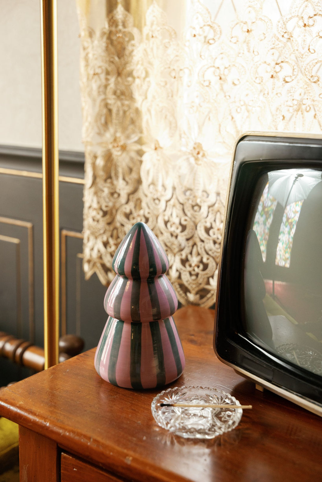 A decorative striped ceramic Christmas tree candle in pink, gray, and black sits on a wooden table next to a vintage television and a clear glass ashtray, with ornate cream lace curtains in the background.