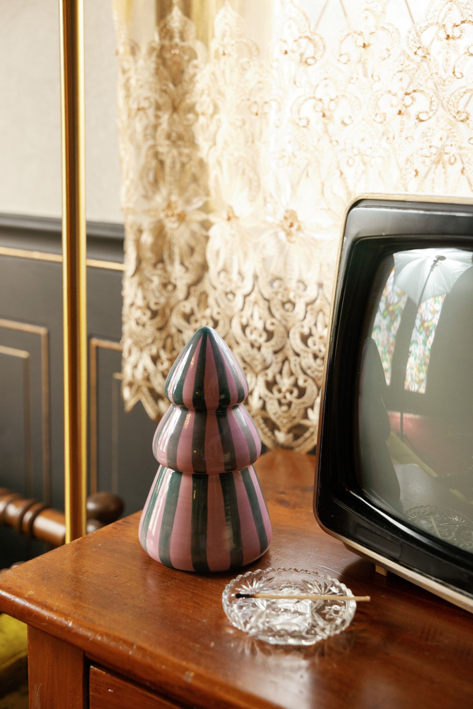 A decorative striped ceramic Christmas tree candle in pink, gray, and black sits on a wooden table next to a vintage television and a clear glass ashtray, with ornate cream lace curtains in the background.