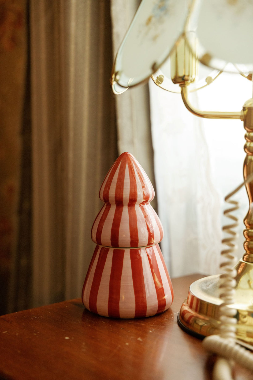 A small decorative ceramic Christmas tree-shaped candle with red and pink vertical stripes sits on a wooden table next to a vintage gold telephone receiver in a softly lit room.
