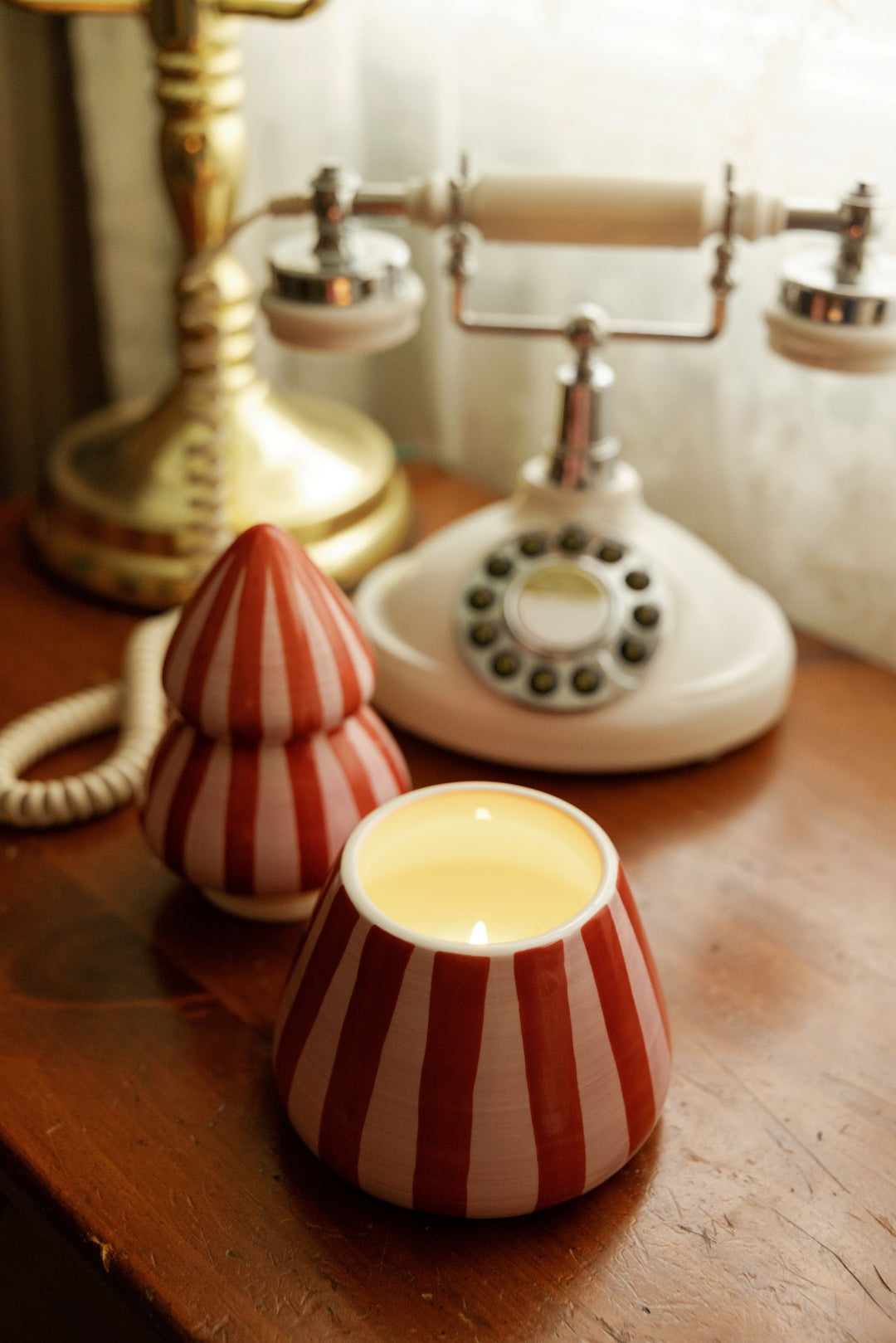 A red and pink vertically striped ceramic candle holder with a lit candle in the foreground, next to a matching striped candle tin, set on a wooden table with a vintage rotary phone and brass lamp in the background.