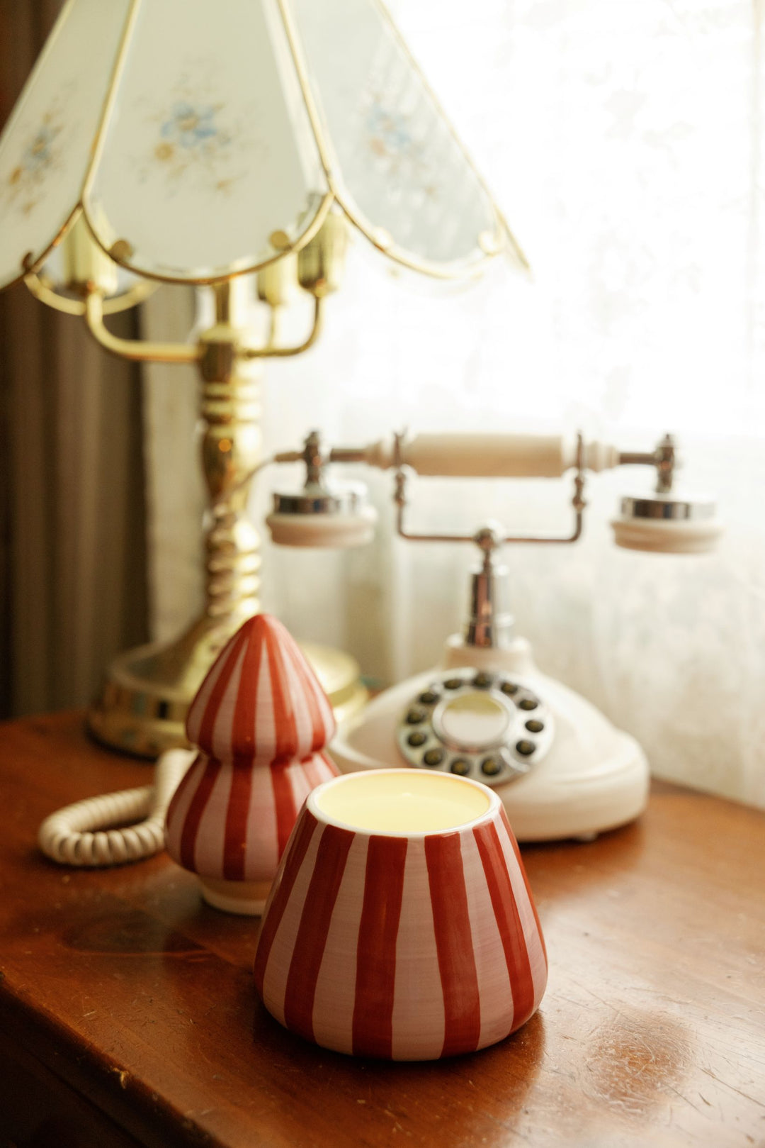 A pair of red and pink vertically striped ceramic candle holders on a wooden table, styled with a vintage cream rotary telephone and a floral table lamp in the background.