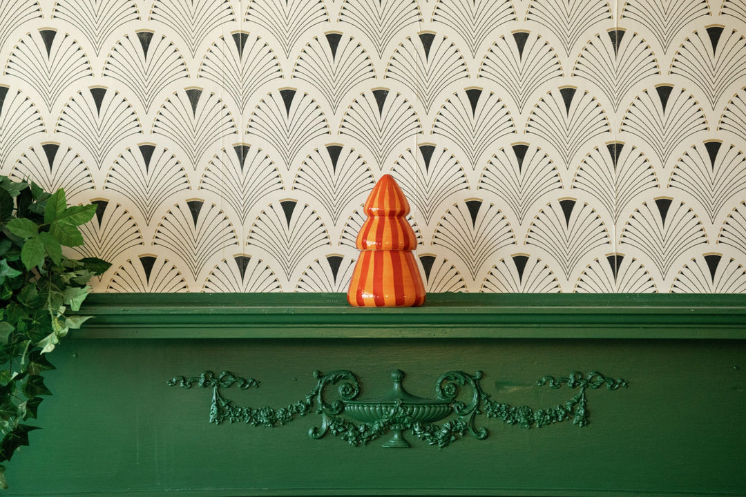A small orange and red striped ceramic Christmas tree candle sits on a dark green mantelpiece against a white wall with a repeating black fan-patterned wallpaper.