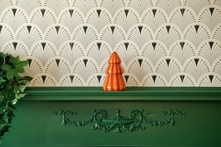 A small orange and red striped ceramic Christmas tree candle sits on a dark green mantelpiece against a white wall with a repeating black fan-patterned wallpaper.