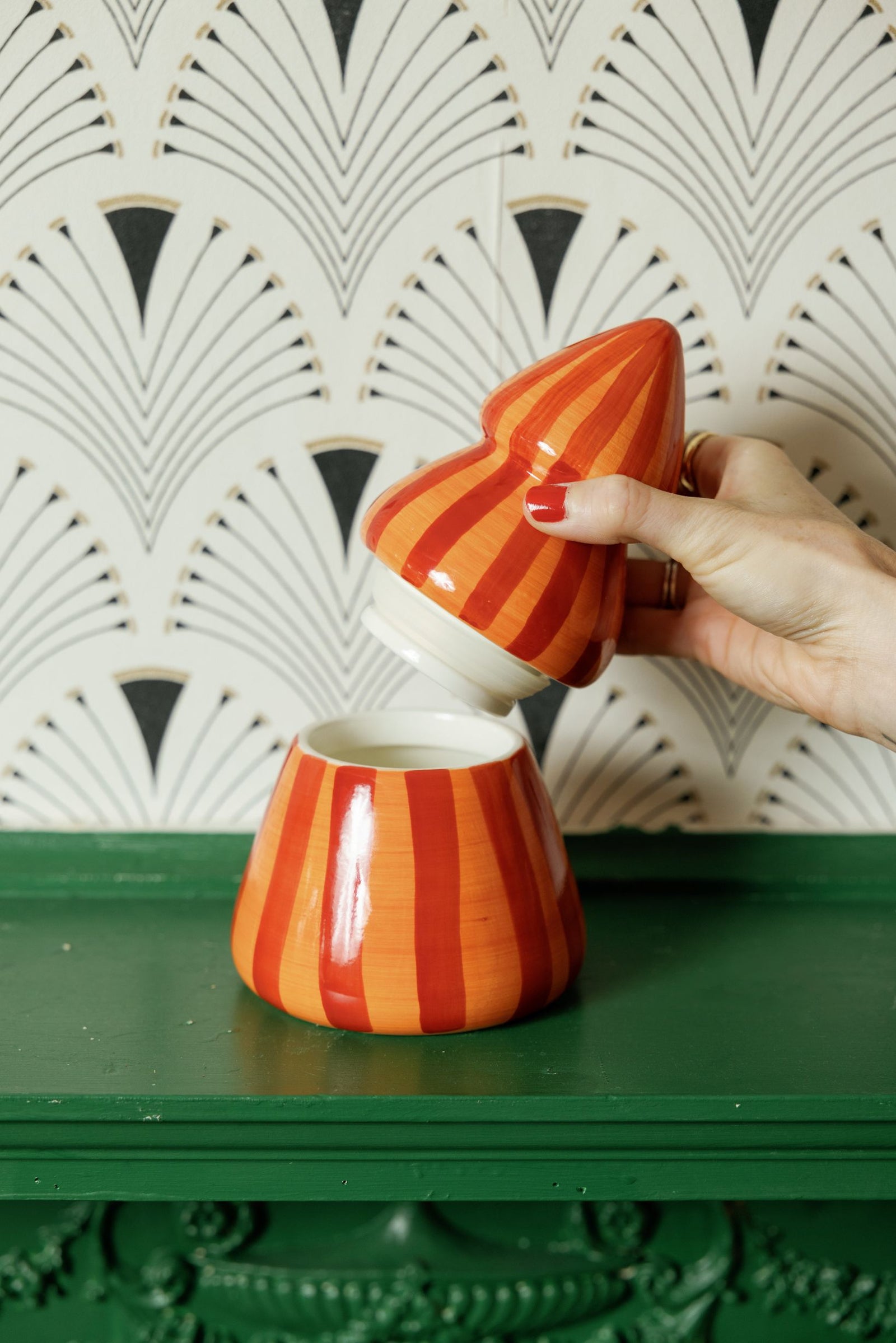 A hand with red nail polish holds a small orange and red striped ceramic candle lid above matching candle on a dark green surface, with patterned wallpaper in the background.