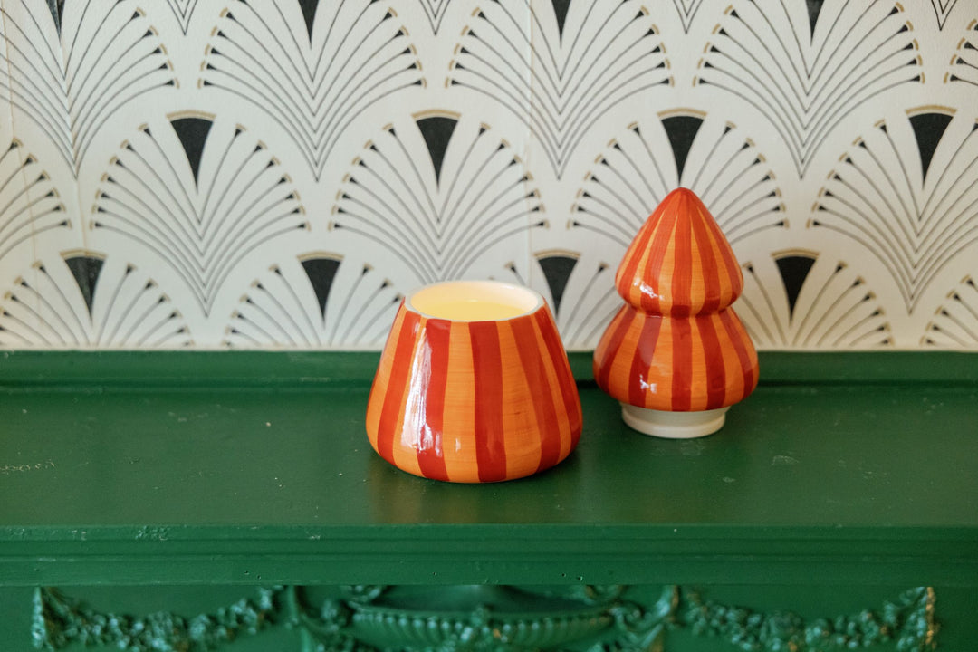 Decorative ceramic candle with lid with orange and red vertical stripes sitting on a dark green mantelpiece in front of a white wall with a black and gray fan pattern.
