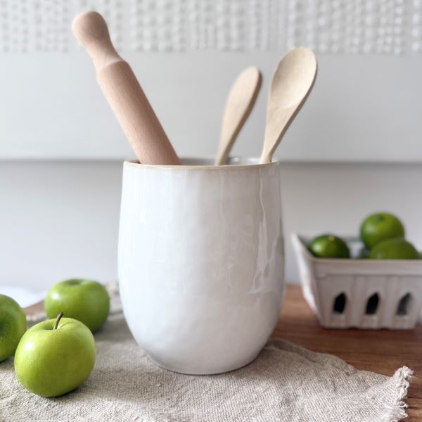 White ceramic utensil pot with wooden spoons on a wooden surface with green apples and a basket in the background.