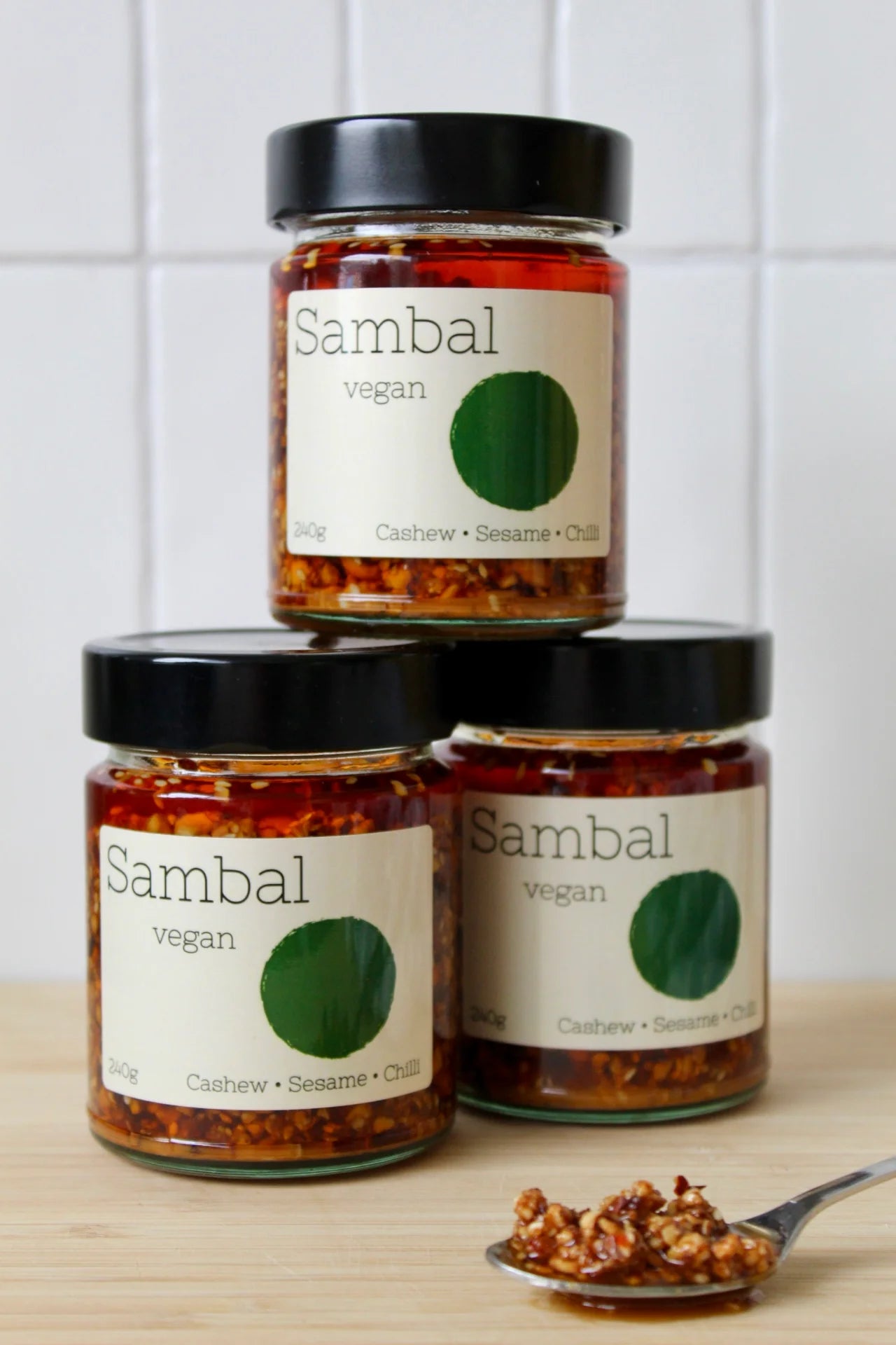 Three jars of Sambal vegan on a wooden surface with a white tiled wall background.
