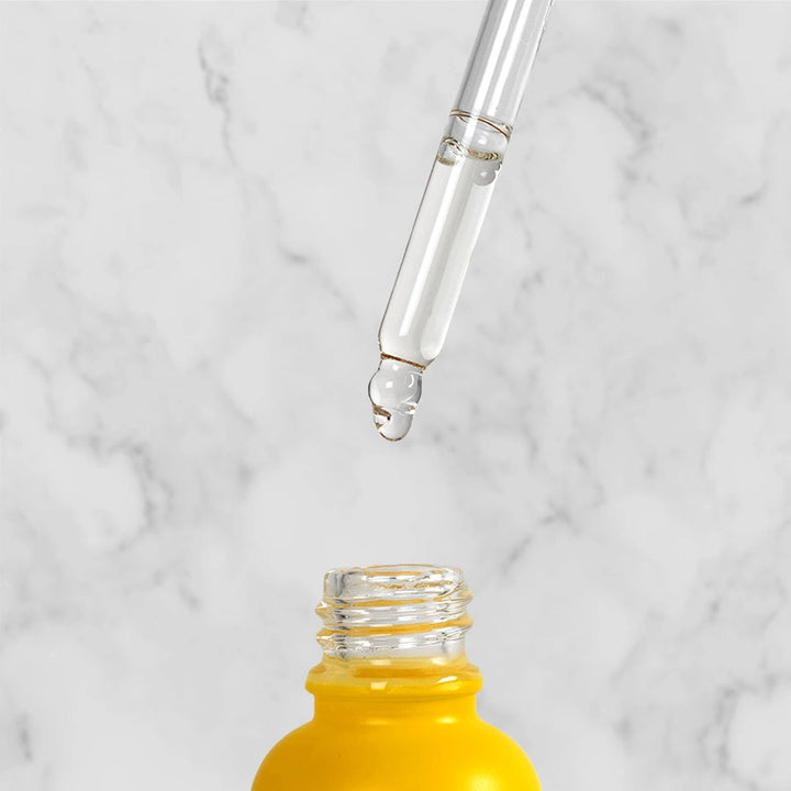 Glass dropper with beard oil dripping into a yellow bottle, in front of a light grey marble background