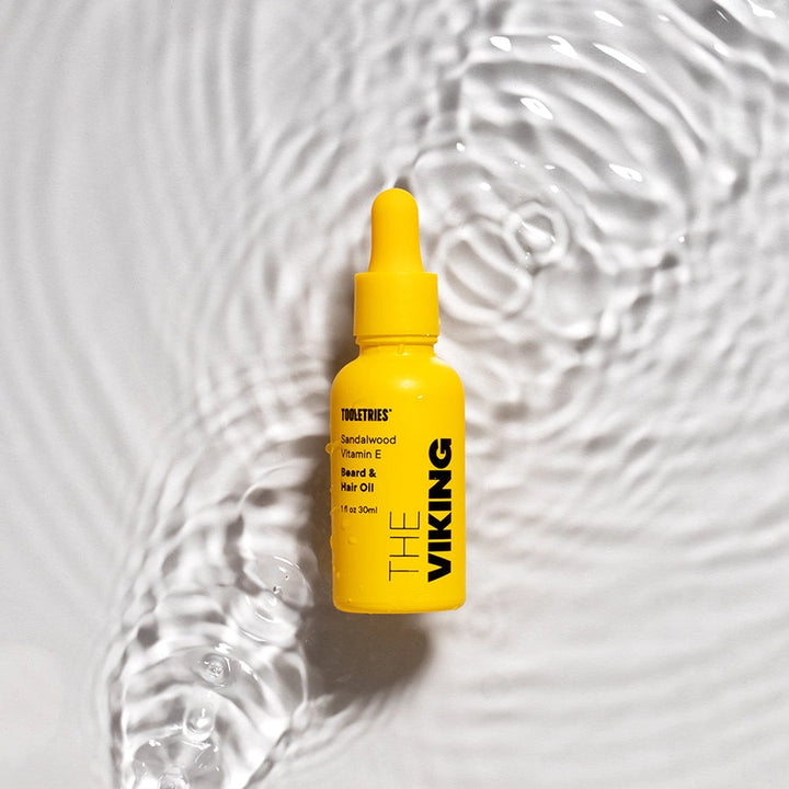A small yellow dropper bottle of The Viking Beard &amp; Hair Oil by Tooletries, set against a textured white background with ripples of clear water.