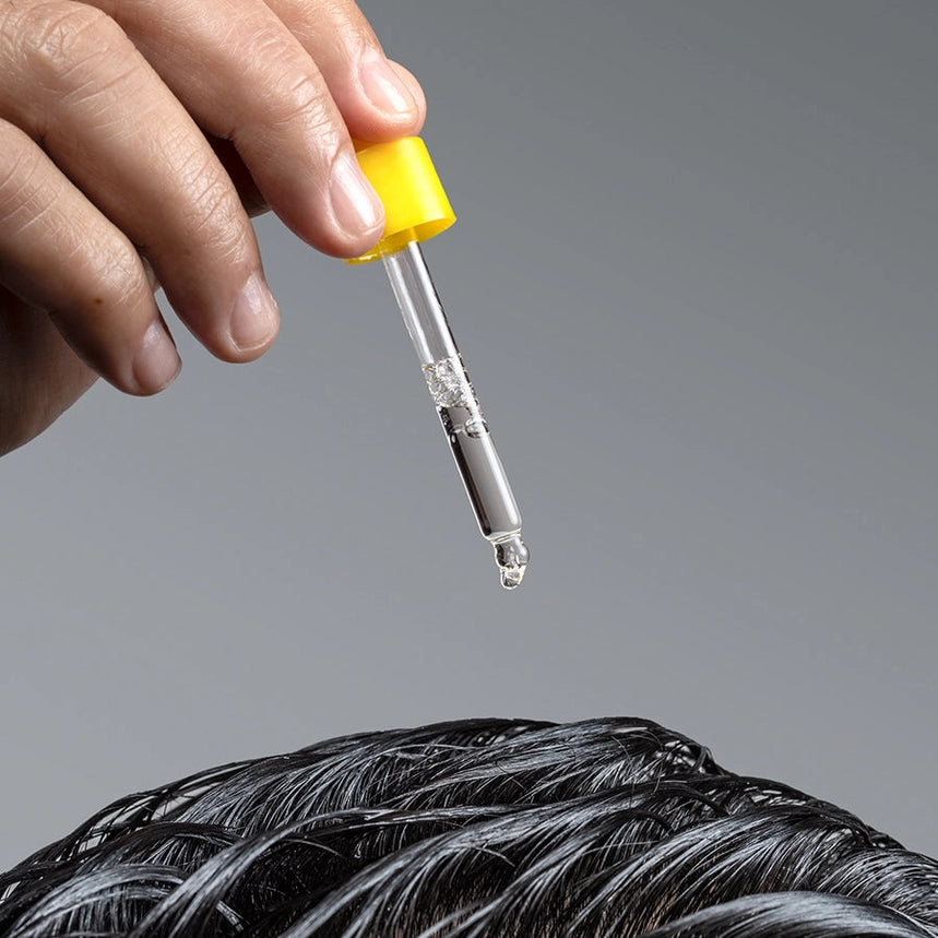 Hand holding a dropper with a clear liquid above hair, on a gray background