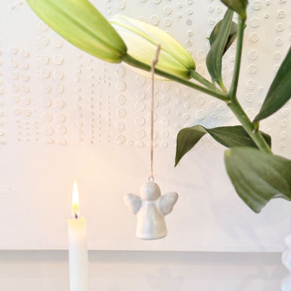 A small white ceramic angel ornament hangs from a string in front of a white textured wall, with a lit white candle at lower left and green lily flower stems at the top right.