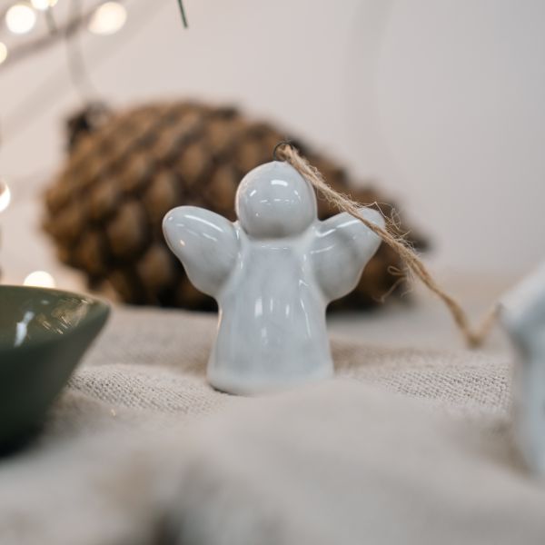 A small glossy white ceramic angel ornament with outstretched wings and a natural jute hanging string, displayed on a textured beige fabric surface with a blurred pinecone and warm string lights in the background.