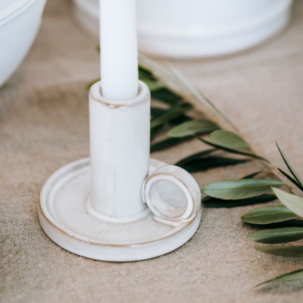 A white ceramic candlestick holder with a matching white candle, set on a beige burlap surface with green leaves in the background.