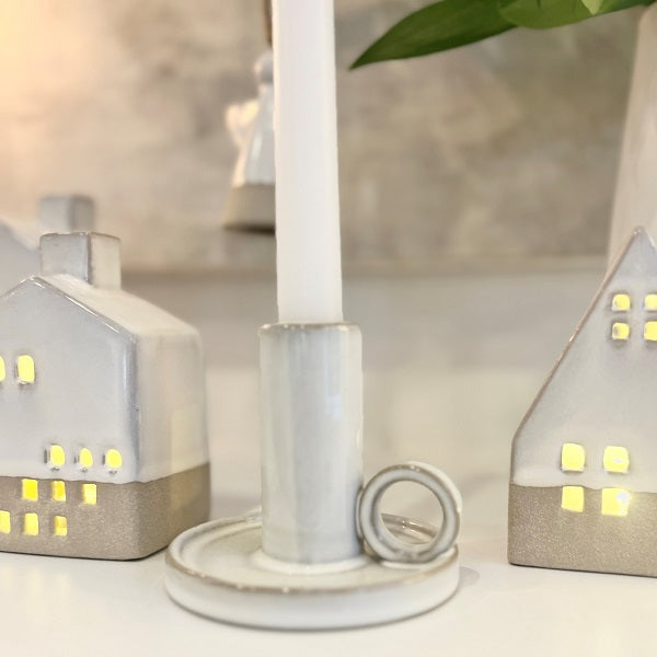 A white ceramic candlestick holder with a matching round base and a small loop on the side, shown with a white candle inserted. The scene is softly lit, with two small house-shaped decorative lights glowing in the background.