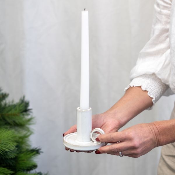 A person holding a white taper candle in a white holder, with their hands and arms visible against a soft light gray backdrop. A green pine branch appears at the lower left edge.