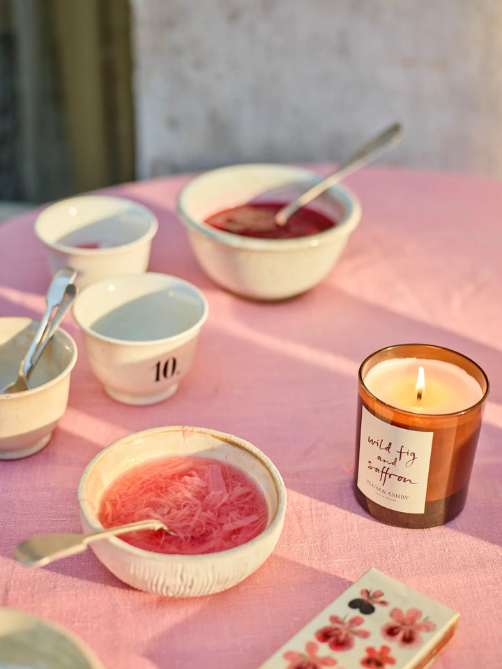 Lit wild fig and saffron candle on a table with pink tablecloth, bowls of jam and condiments