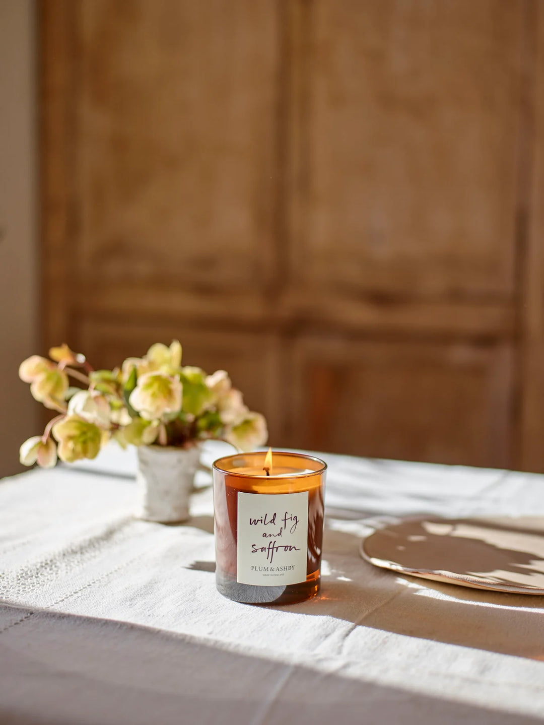 Lit wild fig and saffron candle on a table with a white linen tablecloth and flowers