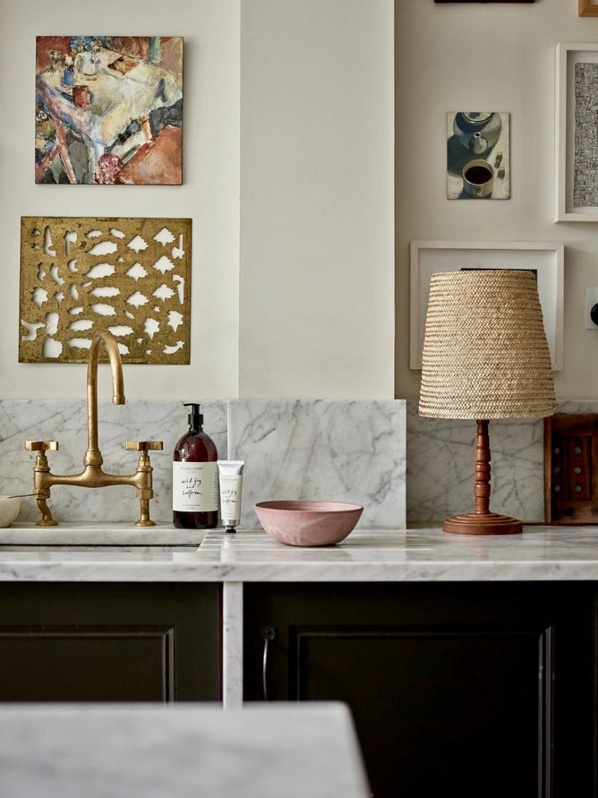 Kitchen bench and sink with lamp, pink bowl, artworks on the wall, and a bottle of wild fig and saffron hand wash and tube of  hand cream