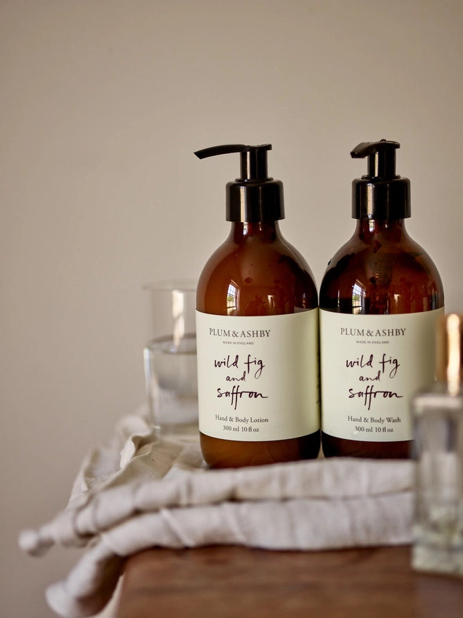 Two amber pump bottles labeled Plum &amp; Ashby Wild Fig and Saffron hand &amp; body lotion and hand &amp; body wash, standing on a draped white cloth on a wooden surface against a warm beige background.