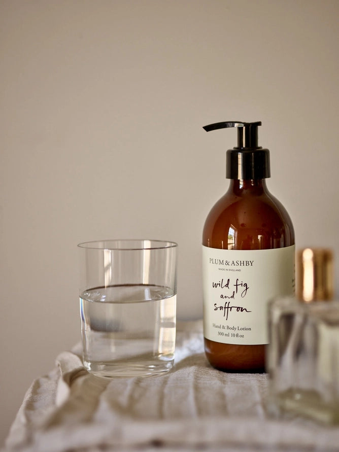 Plum &amp; Ashley wild fig and saffron lotion bottle with a glass of water on a neutral background