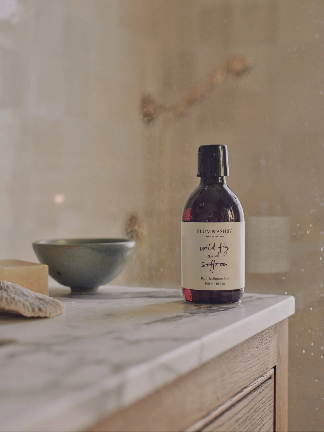 Amber bottle of wild fig and saffron bath and shower gel, on a marble bench in a bathroom with neutral tiles